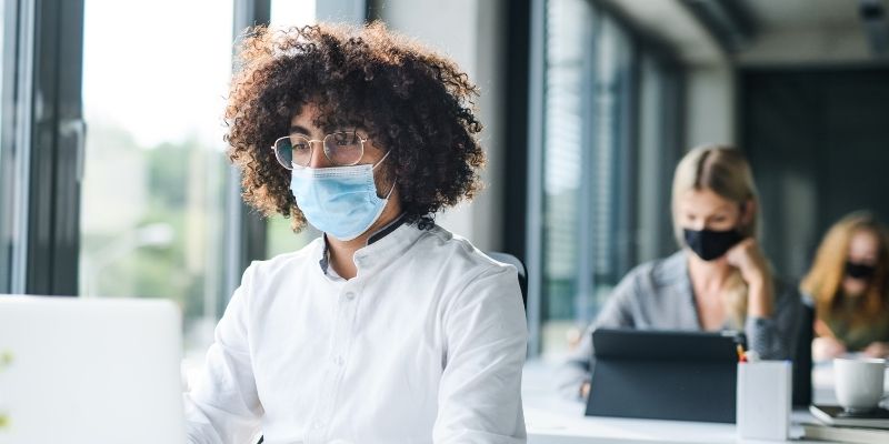 Office workers wearing COVID-19 face coverings or masksworking at desks with safe social distancing