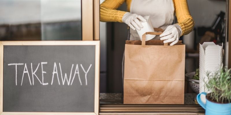 Female restauranteur packing takeaway meal into brown paper bag for pickup or delivery during COVID-19 pandemic